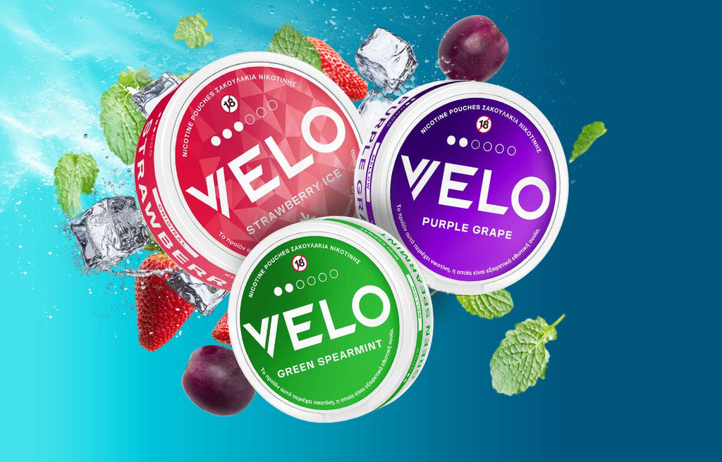 Τρία δοχεία σακουλιών νικοτίνης VELO - Φράουλα Ice (κόκκινο), Μωβ σταφύλι (μοβ) και Πράσινη μέντα (πράσινο) - με φράουλες, σταφύλια, φύλλα μέντας και παγάκια σε μπλε φόντο.