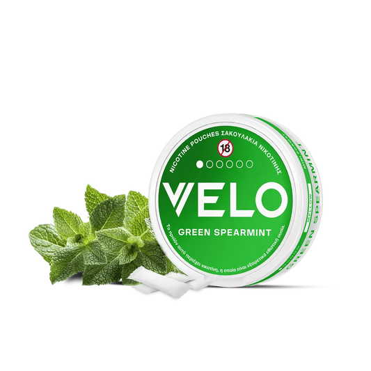 Ένα στρογγυλό λευκό και πράσινο δοχείο VELO Green Spearmint είναι τοποθετημένο δίπλα σε φρέσκα φύλλα μέντας, με διάφορα φυτικά σακουλάκια νικοτίνης διάσπαρτα μπροστά.