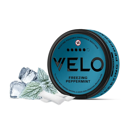 Ένα μπλε κουτάκι VELO Freezing Peppermint βρίσκεται δίπλα σε παγάκια, φύλλα μέντας και δύο λευκά σακουλάκια νικοτίνης σε λευκό φόντο, τονίζοντας τη δροσιστική γεύση μενθόλης του προϊόντος.
